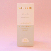 On a pink background is a neutral colored bar of chocolate with gold foiled text that reads, "Valerie rice & sesame Milk Chocolate".