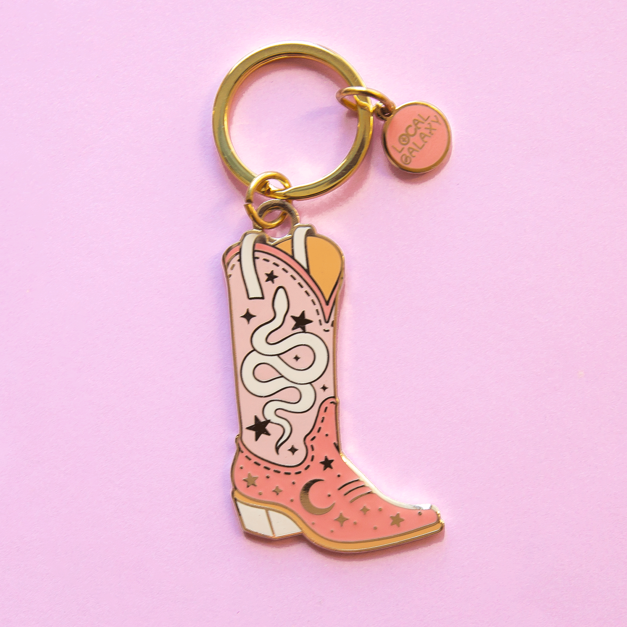 On a pink background is a pink and ivory cowgirl boot shaped keychain with a gold loop for hanging. 