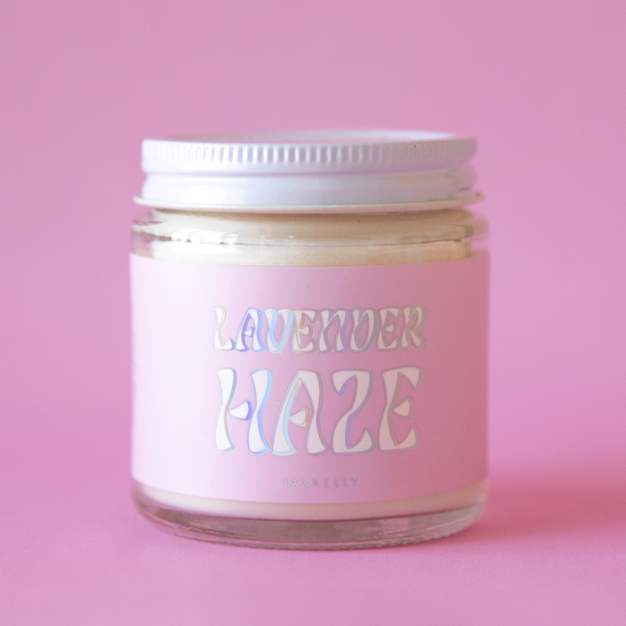 On a pink background is a 4oz glass jar candle with a light pink label that reads, &quot;Lavender Haze&quot; in a holographic wavy font.