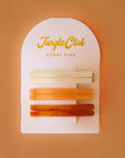 On a brown background is an arched packaging with embossed text at the top that reads, "Jungle Club Bobby Pins" along with six thin hair clips in three colors from an ivory shade to orange to a honey rust color.