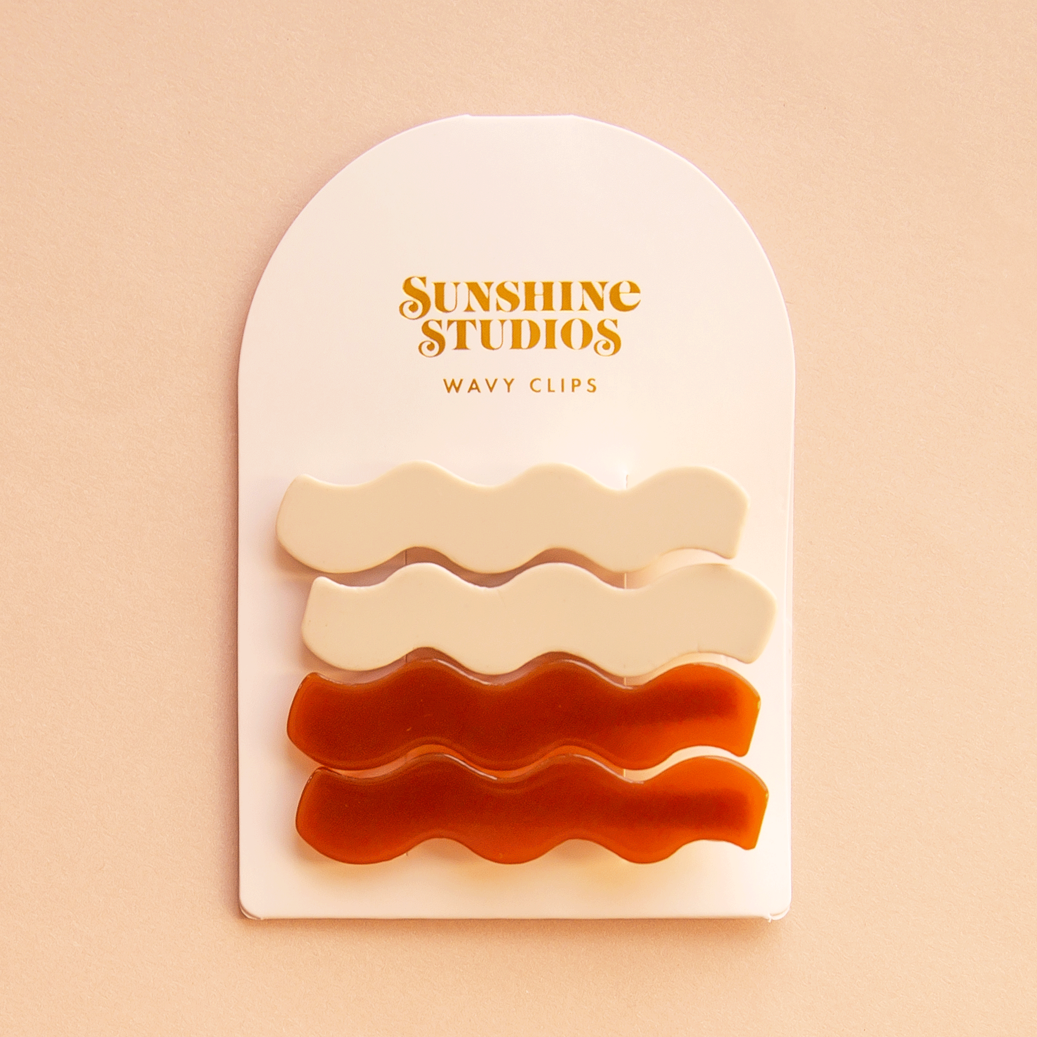 On a peach background is two pairs of wavy hair clips on a white arched cardboard backing with text at the top that reads, &quot;Sunshine Studios Wavy Clips&quot;.