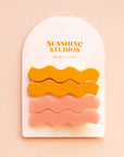 On a pink background two sets of wavy hair clips in a mango orange shade and a light pink shade clipped on a white arched piece of cardboard packaging with orange text at the top that reads, "Sunshine Studios".