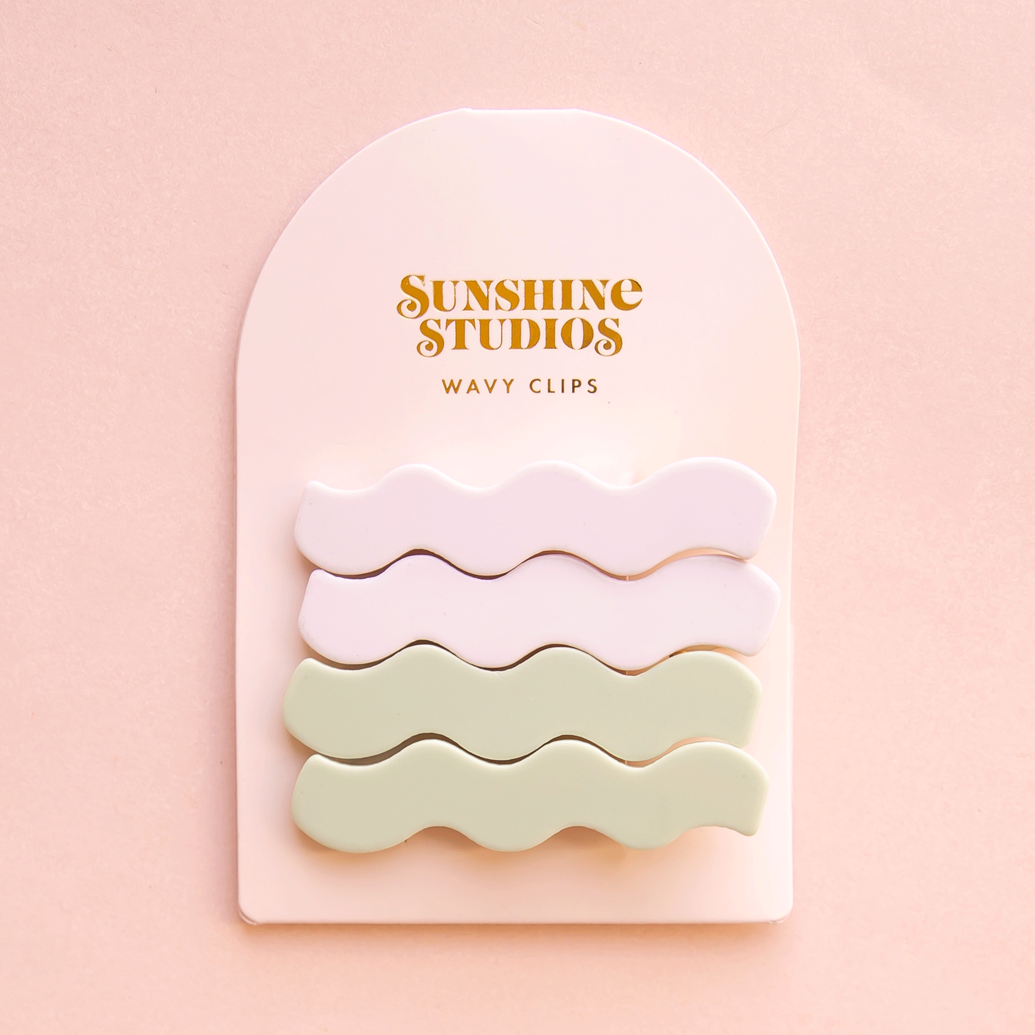 On a pink background is two sets of wavy shaped hair clips in a light green/blue shade and shade of white on an arched packaging with text along the top that reads, &quot;Sunshine Studios&quot;.
