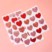 On a pink background is four sheets of heart shaped glitter stickers in shades of red and pink. Edit alt text