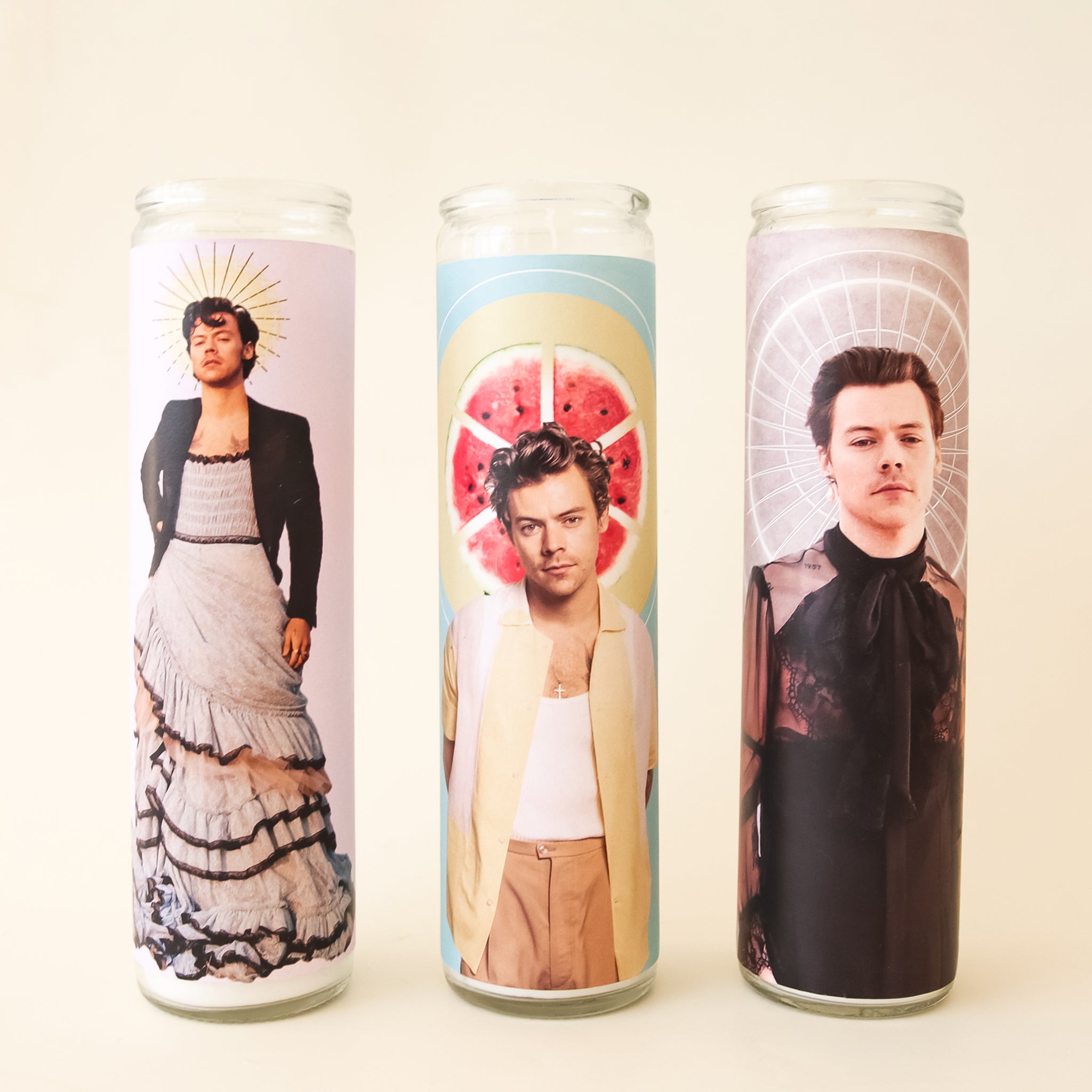 On a white background is a prayer candle with Harry Styles in a dress on the front photographed next to other available Harry Styles prayer candles on our site.