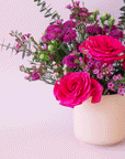 On a purple background is a light pink glazed ceramic planter filled with a pink, purple and green flower arrangement that is not included with purchase.