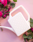 On a pink background is a white watering can with a long spout and a breeze block design on the side.