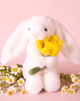 On a pink background is a fuzzy white bunny stuffed animal toy with long floppy ears and holding a stuffed daffodil.