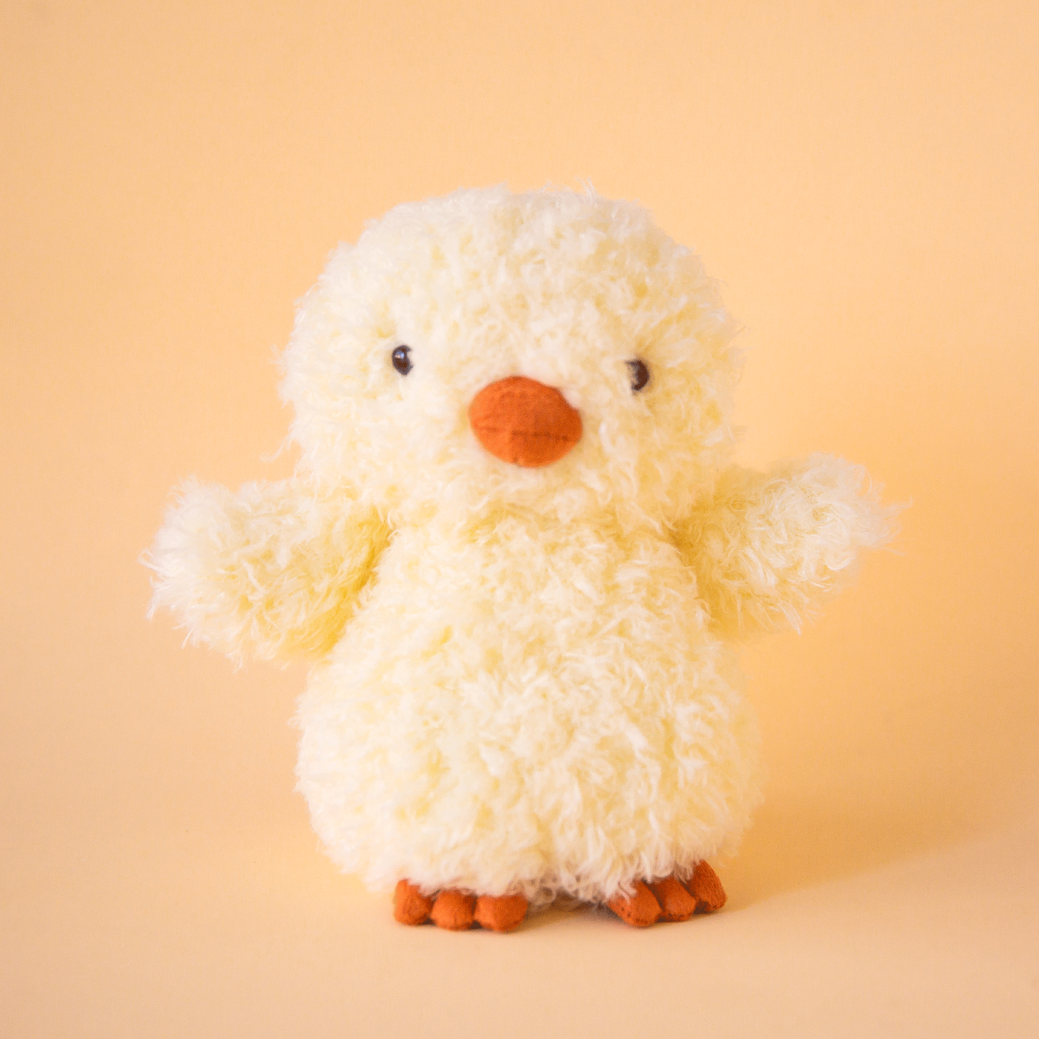 On a yellow background is a super light yellow fuzzy chick stuffed animal toy with an orange beak and feet.