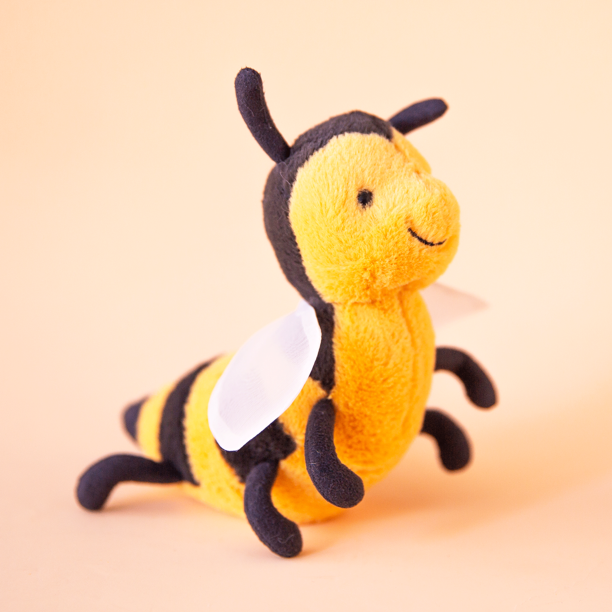 On a peachy background is a yellow and black bee stuffed animal with a smiling face, a fuzzy body and round wings.