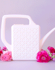 On a purple background is a white watering can with a long spout and a breeze block design on the side.