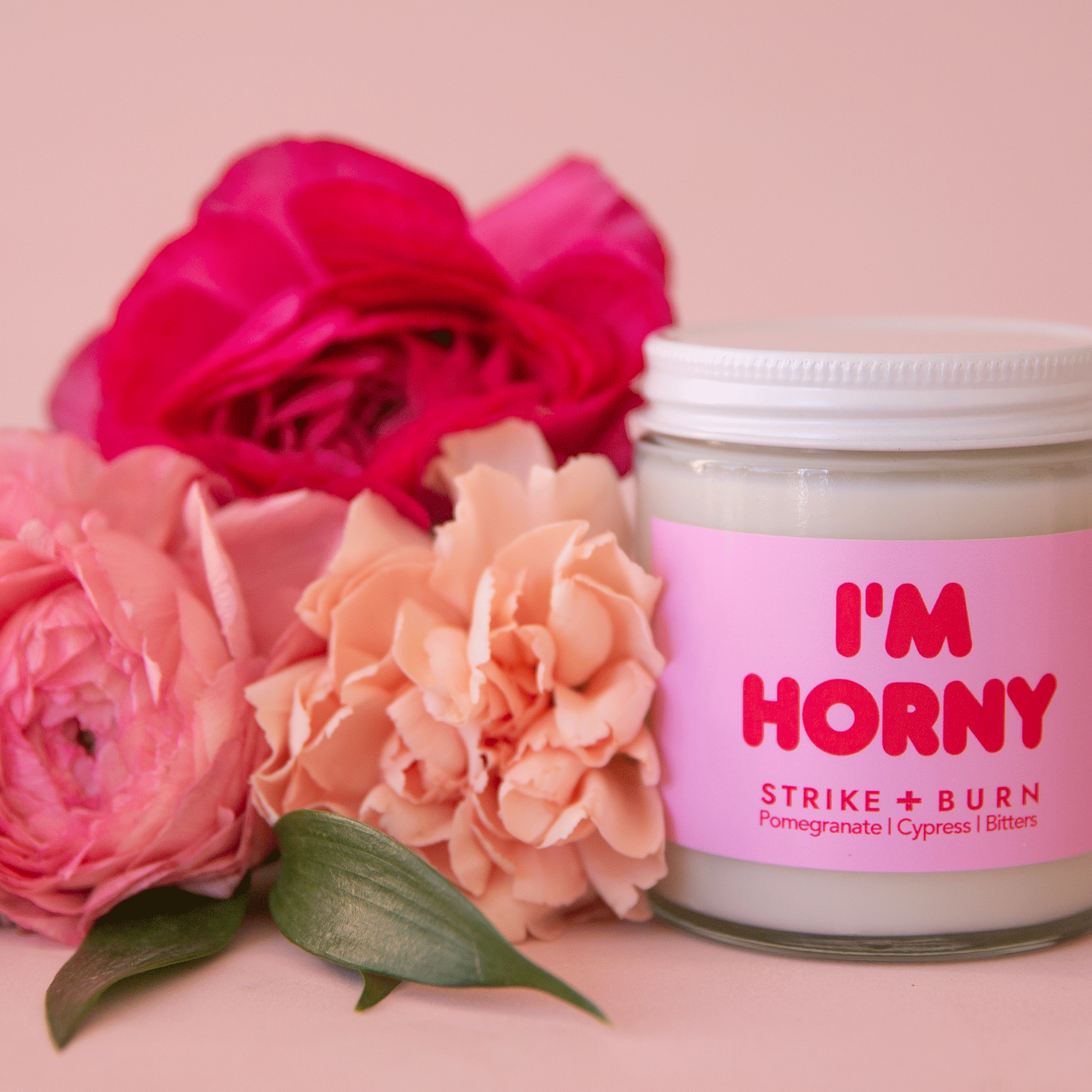 On a pink background is a glass candle jar with a pink and red label that reads, &quot;I&#39;m Horny&quot;.