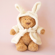 On a pink background is a brown stuffed animal bear wearing a fuzzy white rope with bunny ears.