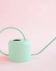On a pink background is a light blue watering can with a long spout and a rounded handle.