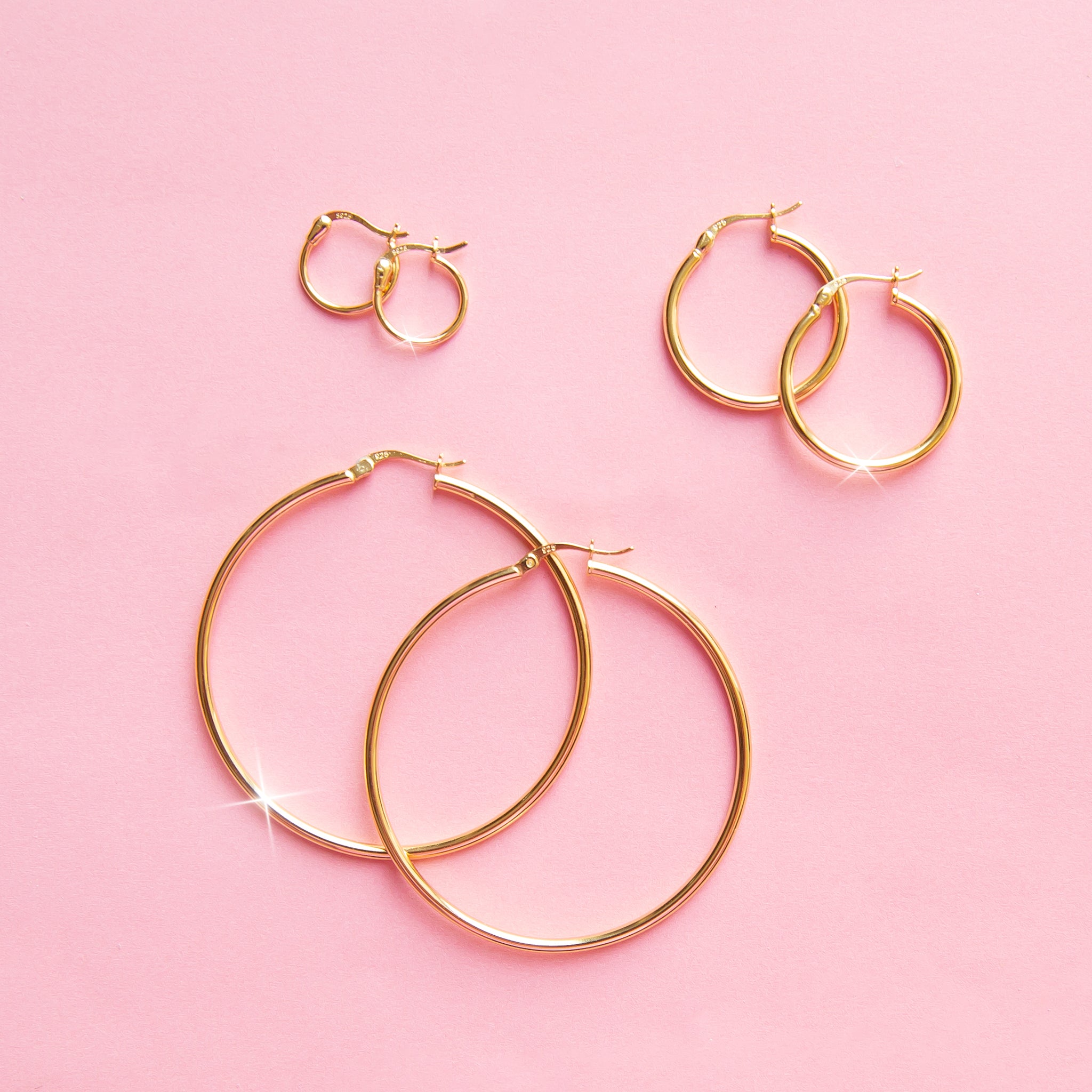 On a pink background is three different sized pairs of thin hoop earrings. 