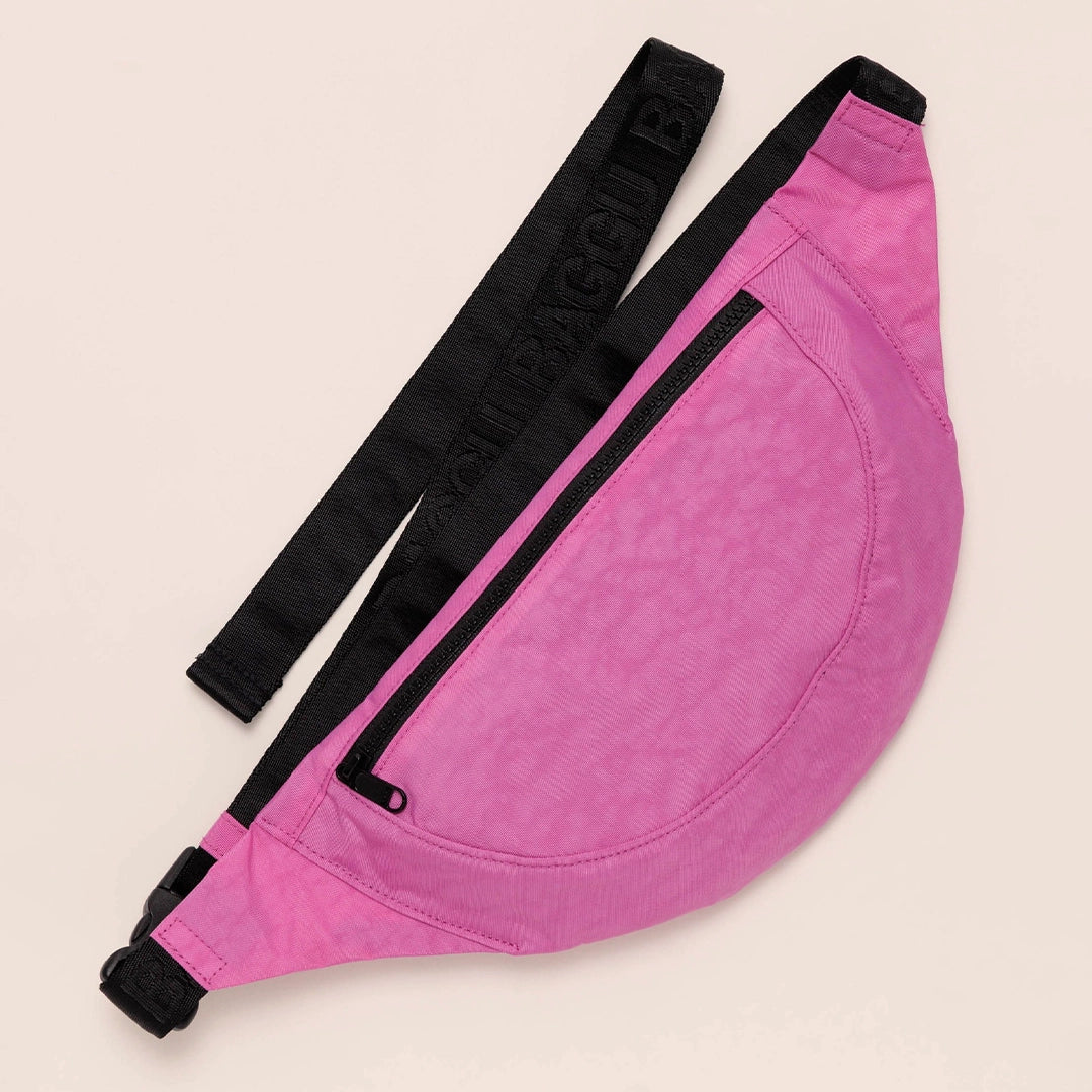 On a tan background is a crossbody fanny pack in a bright pink shade with black details and an outside zipper.