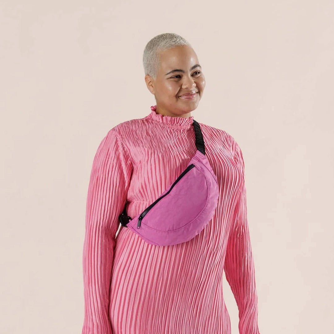 On a light pink background is a model wearing a crossbody fanny pack in a bright pink shade with black details and an outside zipper. 
