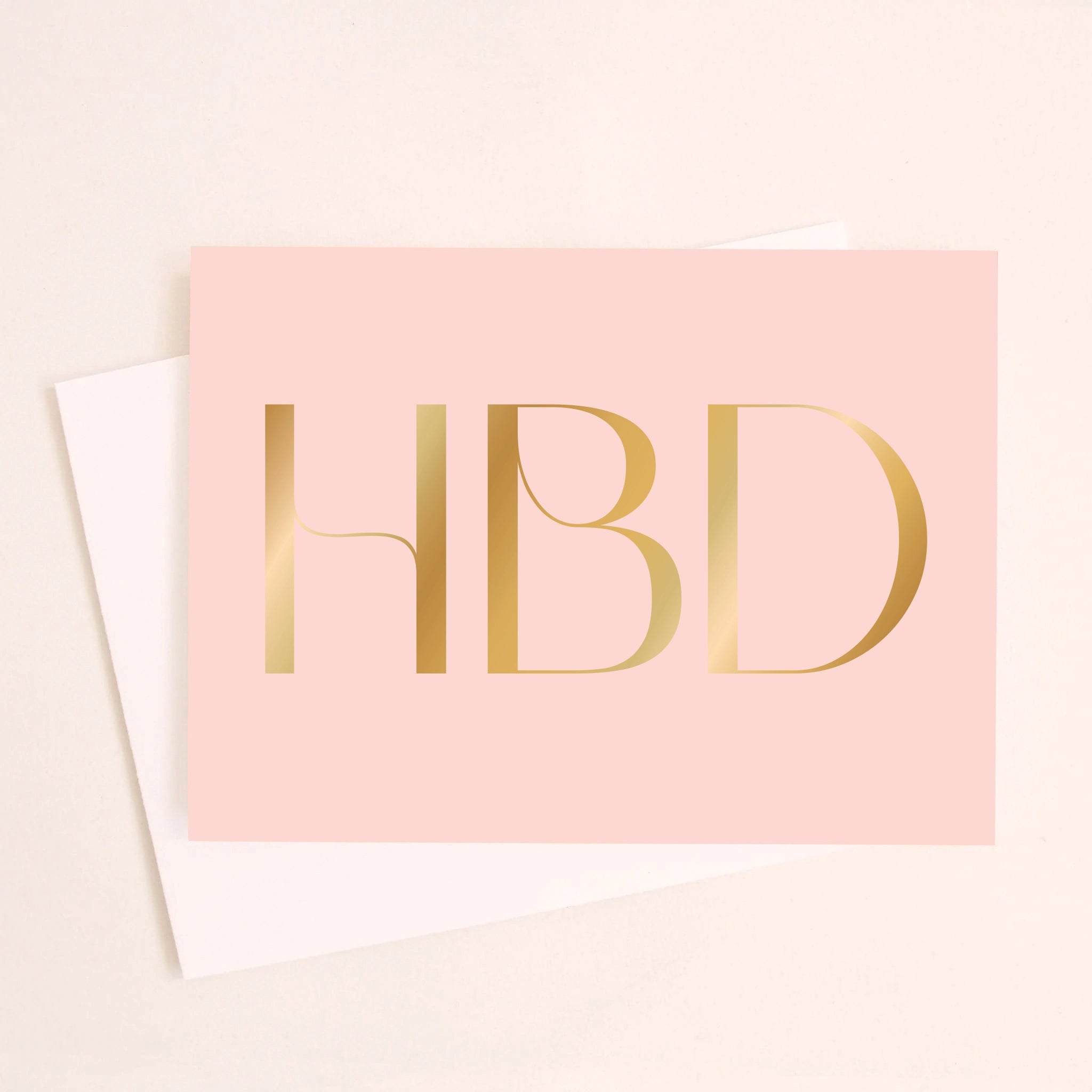 On an ivory background is a light pink greeting card with large gold foil text in the center that reads, &quot;HBD&quot; in all caps. Also included is a white envelope.