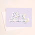 On an ivory background is a light purple/blue greeting card with white / holographic outlined text that reads, "Happy Birthday" along with a white envelope.