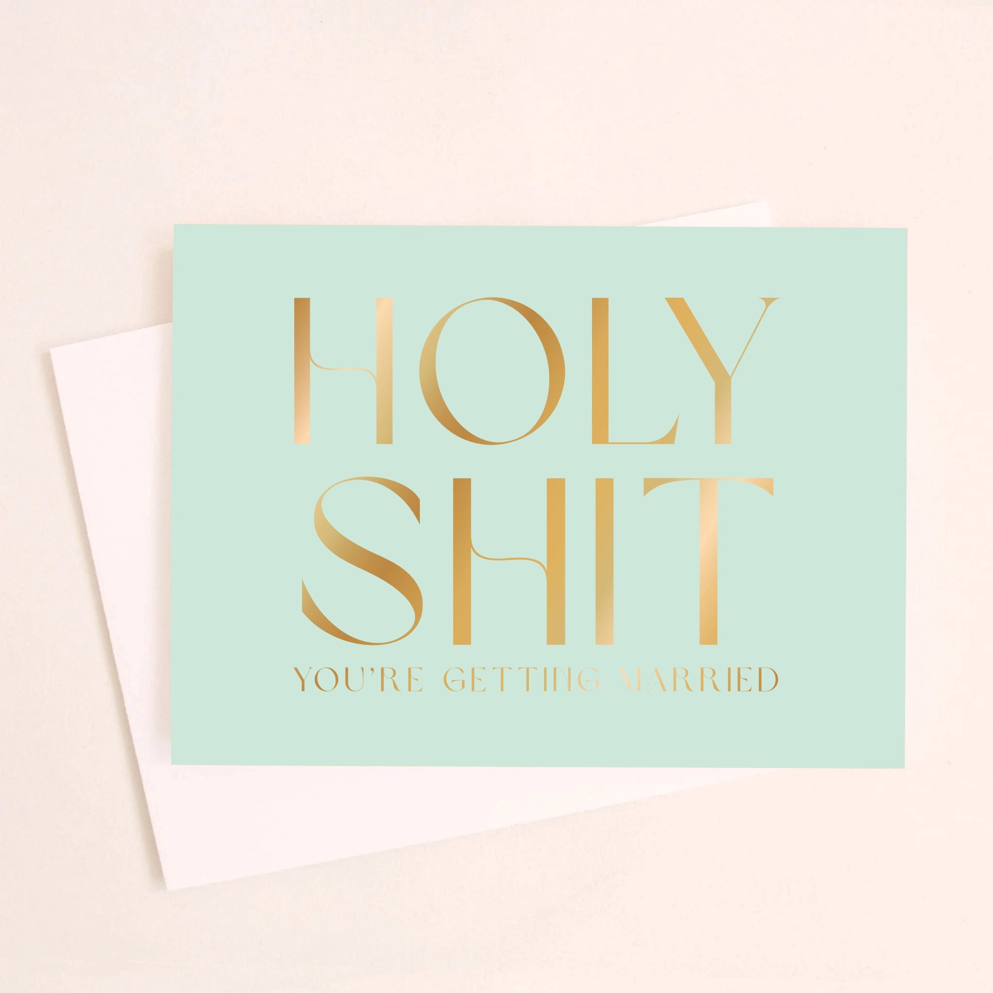 On an ivory background is a teal greeting card that reads, &quot;Holy Shit You&#39;re Getting Married&quot; in gold foil letters and a matching envelope beside it.