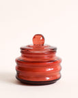 Red colored translucent beam candle vessel with white wax candle, and matching glass lid.