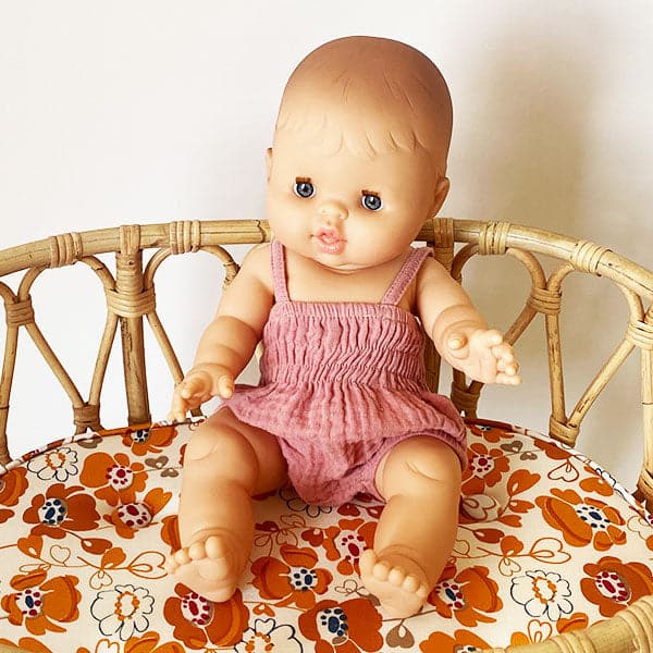 A European baby girl doll wearing a pink romper and sitting in a rattan crib in front of a cream background.