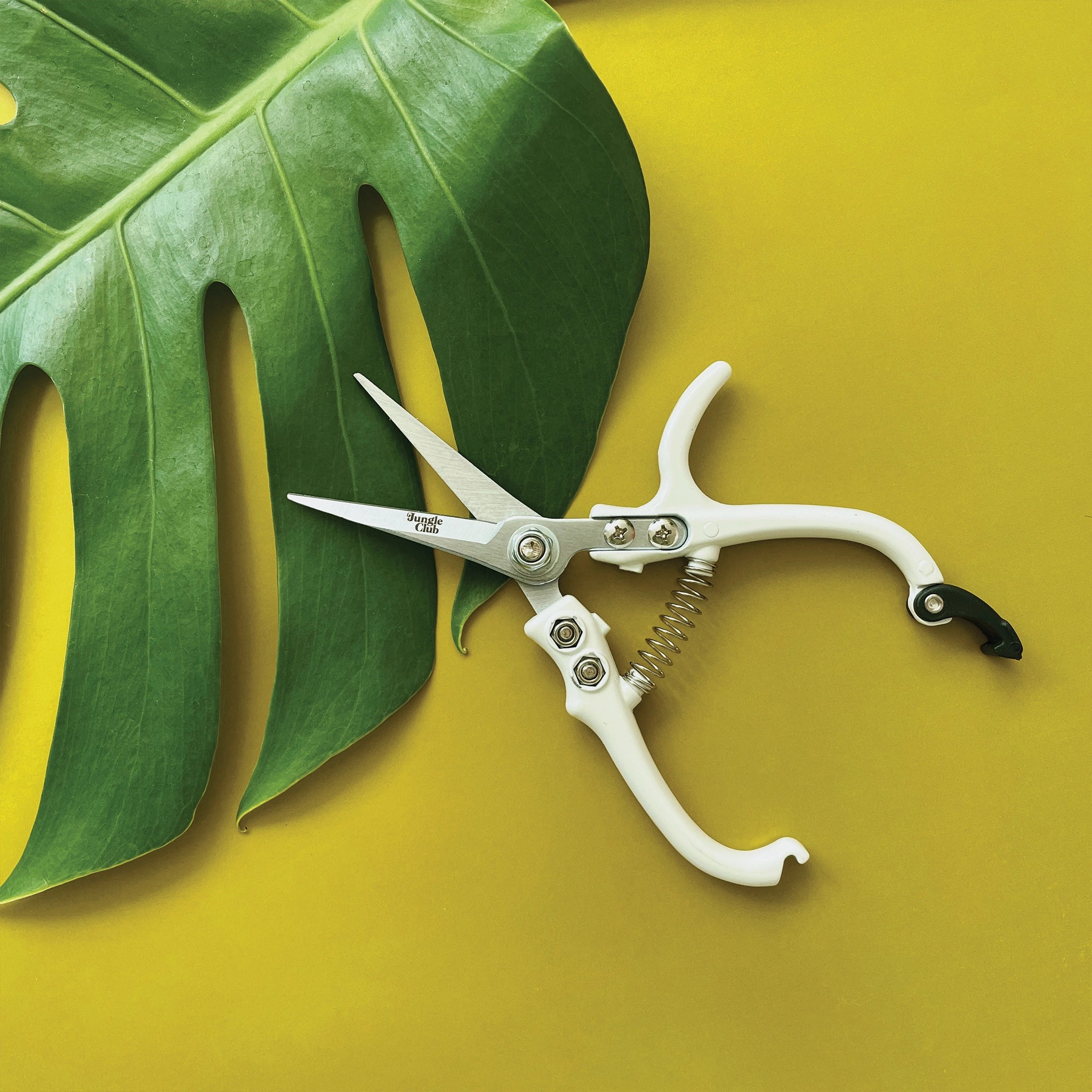 Pair of pruning shears with white handles and metal blades. The outer blade reads &#39;jungle club&#39; in small, playful lettering. The pruning shears have a black clasp at the top of the handles to secure them closed.