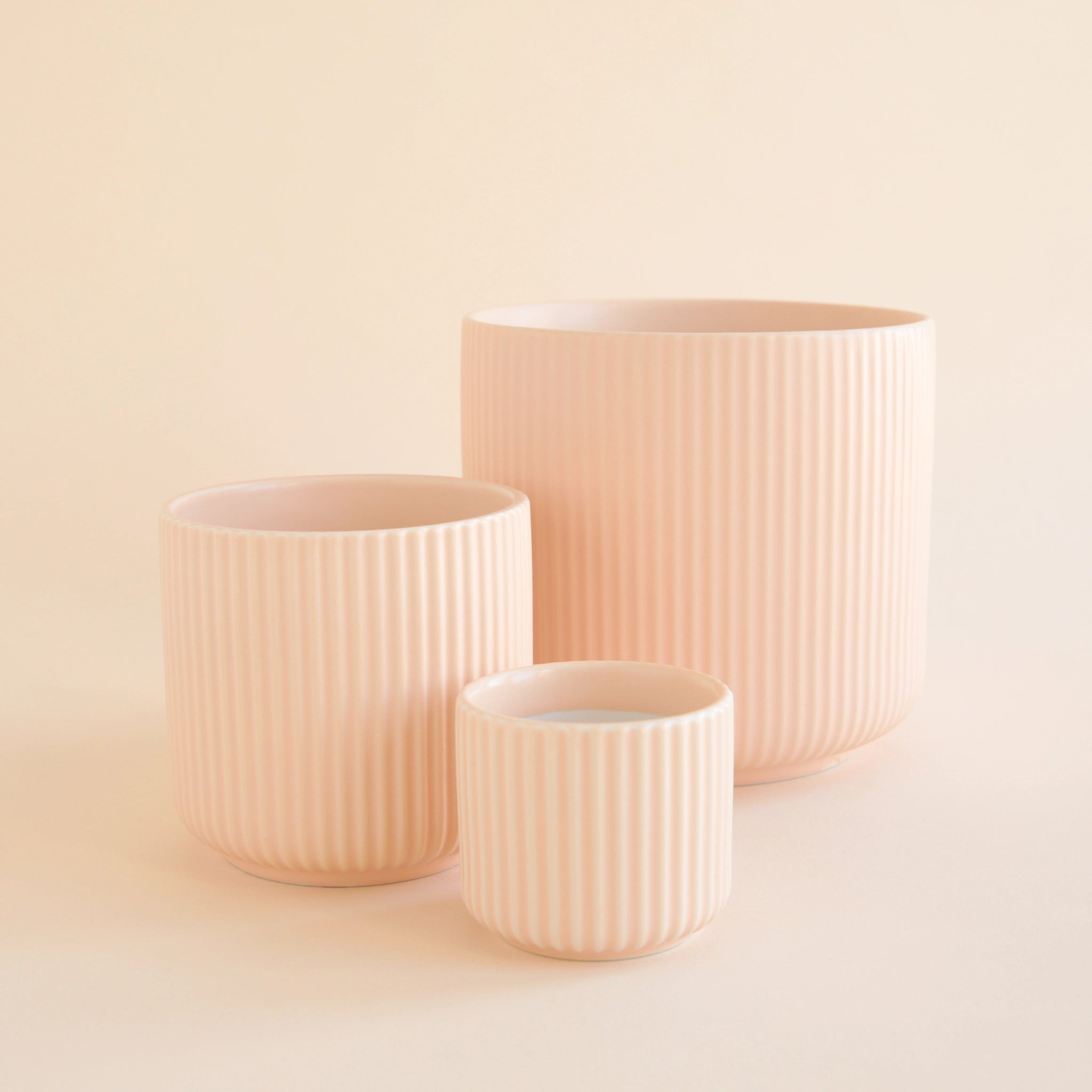 On an ivory background is three different sized pink ceramic planters with fluting and a rounded base.
