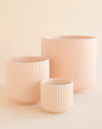 On an ivory background is three different sized pink ceramic planters with fluting and a rounded base.