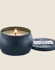 On a cream background is a round tin single wick candle in a navy blue shade along with a label on the lid that reads, "Voluspa Makassar Ebony & Peach".