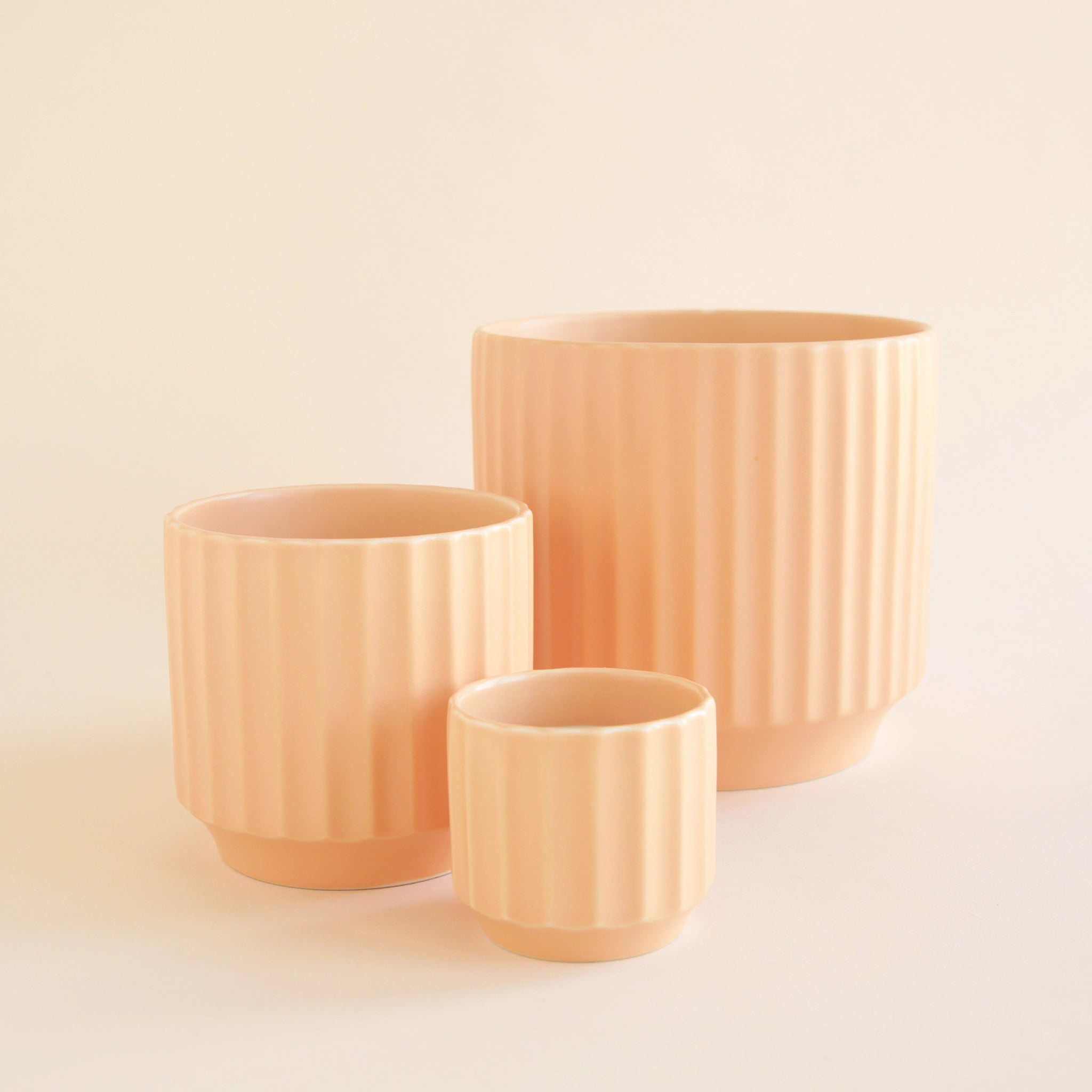 On an ivory background is three different shaped ceramic pots in a warm toned pink shade.
