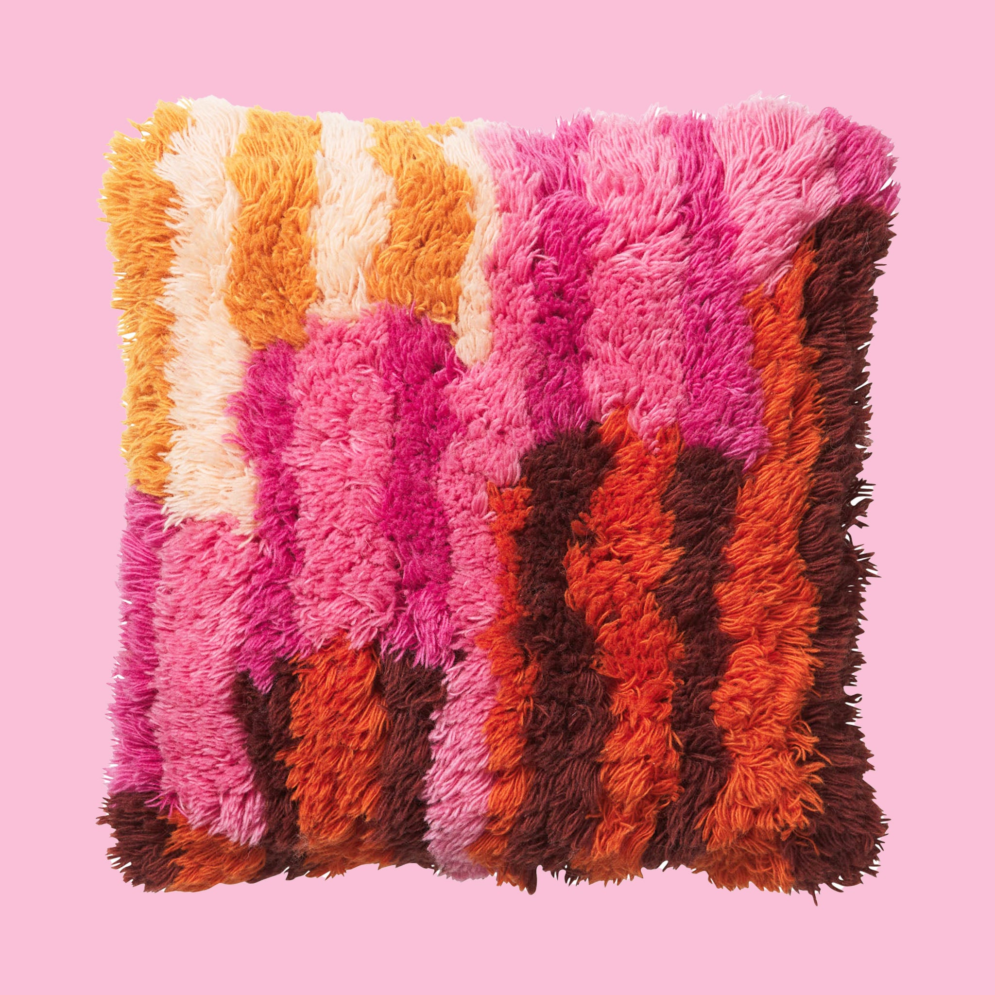 On a pink background is a multi colored shag pillow in orange, brown, pink and yellow shades. 
