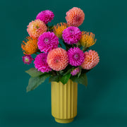 A beautiful chartreuse fluted cylindrical vase sits in middle of a page. Inside the vase are beautiful Dahlia flowers- orange, pink and coral. There are green leaves spilling out of the vase. The flowers and vase are on a beautiful aquamarine colored background.