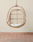 On a neutral background is a natural rattan egg shaped hanging chair with an ivory cushion and rope details.