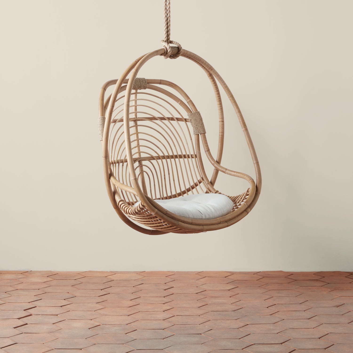 On a neutral background is a natural rattan egg shaped hanging chair with an ivory cushion and rope details. 
