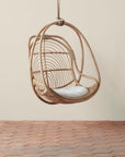 On a neutral background is a natural rattan egg shaped hanging chair with an ivory cushion and rope details.