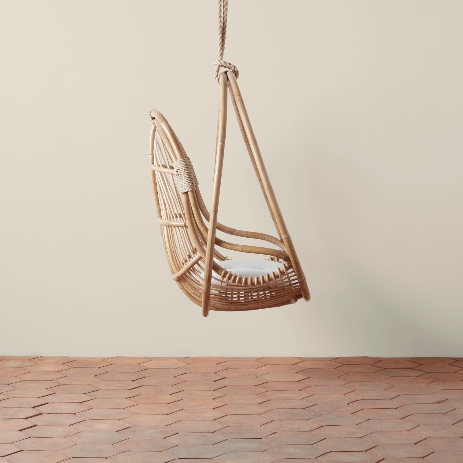 On a neutral background is a natural rattan egg shaped hanging chair with an ivory cushion and rope details.