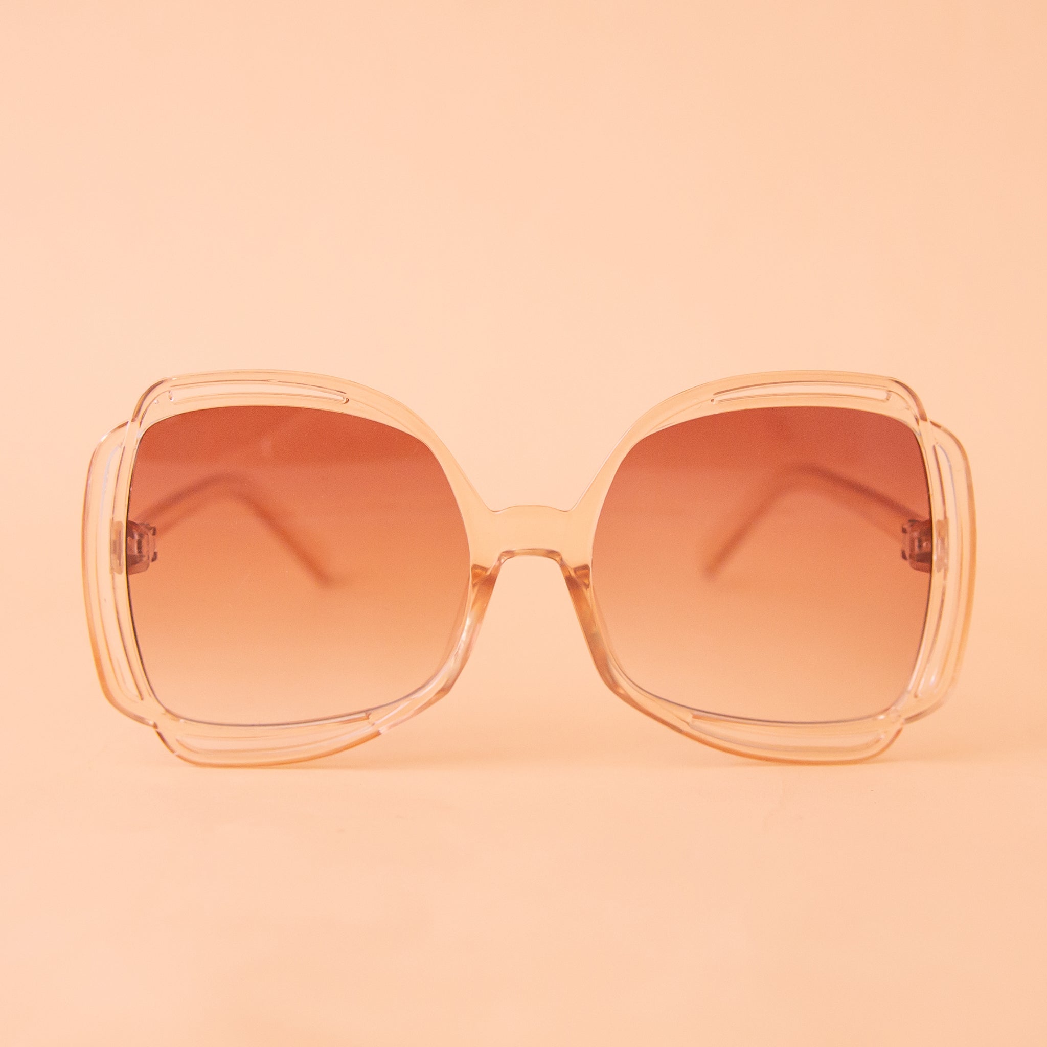A pair of sunglasses with a clear peachy frame and pinkish gradient lenses.
