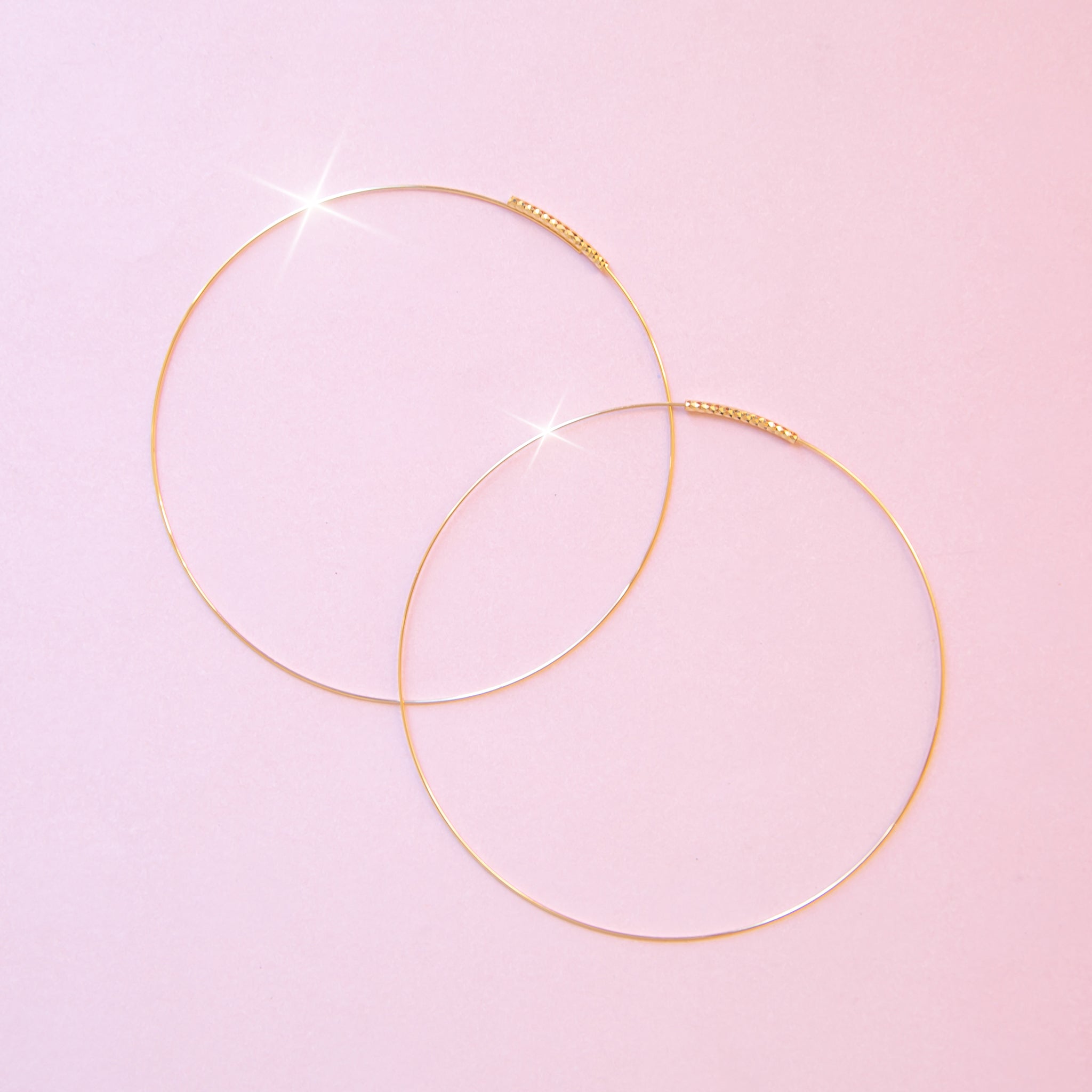On a pink background is a pair of dainty gold hoop earrings. 