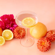 On a pink background is a clear coupe glass with a beverage inside.