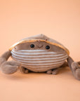 On a tan background is a grey ish brown crab stuffed toy with a sweet smiling face.