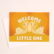 On an ivory background is an orange greeting card with an orange gradient heart background and two snails facing one another with a baby snail on the back of one along with text above and below that reads, "Welcome Little One".