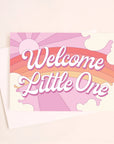 On a light pink background is a greeting card featuring a rainbow and sun graphic and white text in the center that reads, "Welcome Little One".
