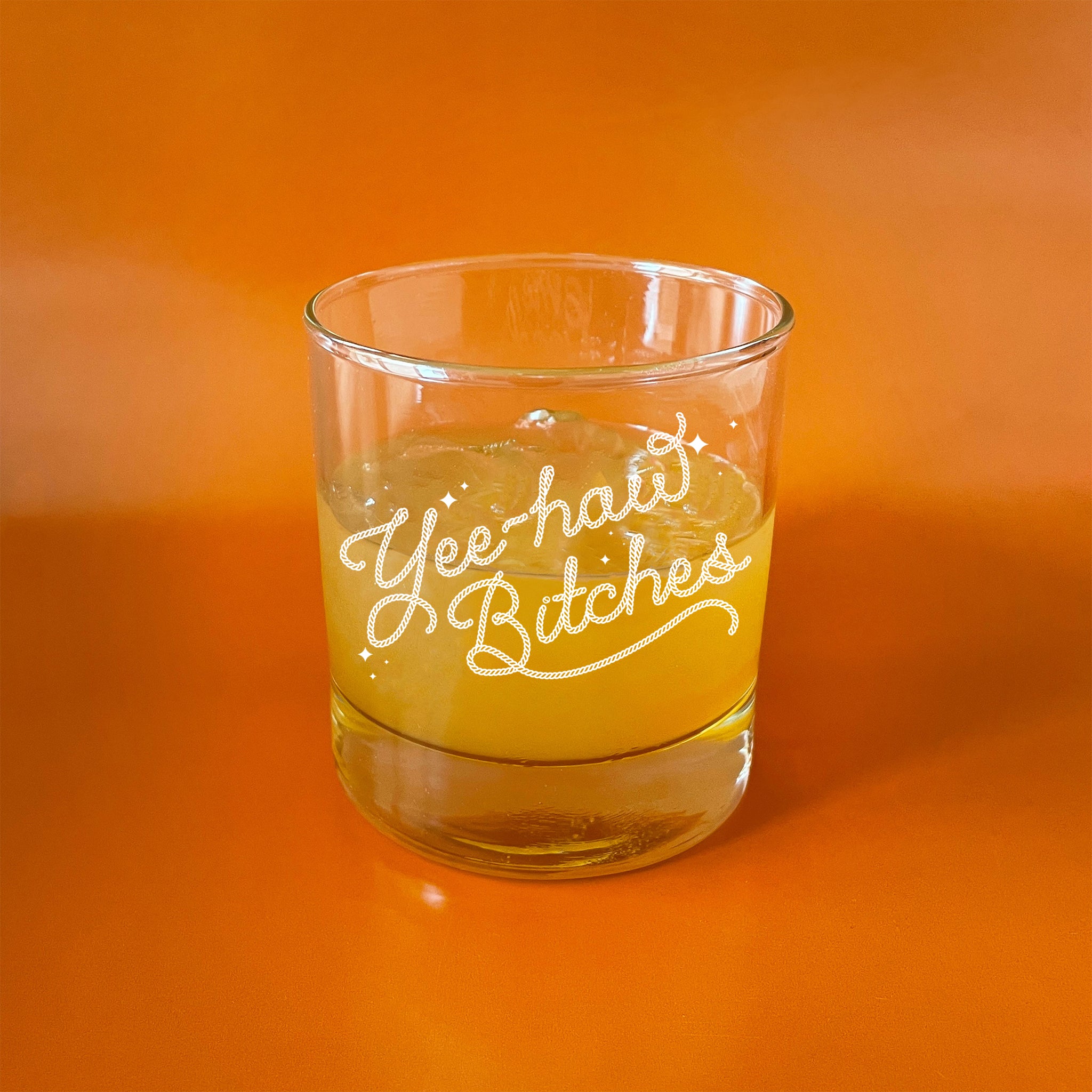 On an orange background is a short glass tumbler with white screenprinted text that reads, &quot;Yee-haw Bitches&quot;.