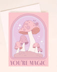 On a light pink background is a pink and purple card with an arch and mushroom design as well as text along the bottom that reads, "You're Magic". A white envelope is included with purchase, also shown here.
