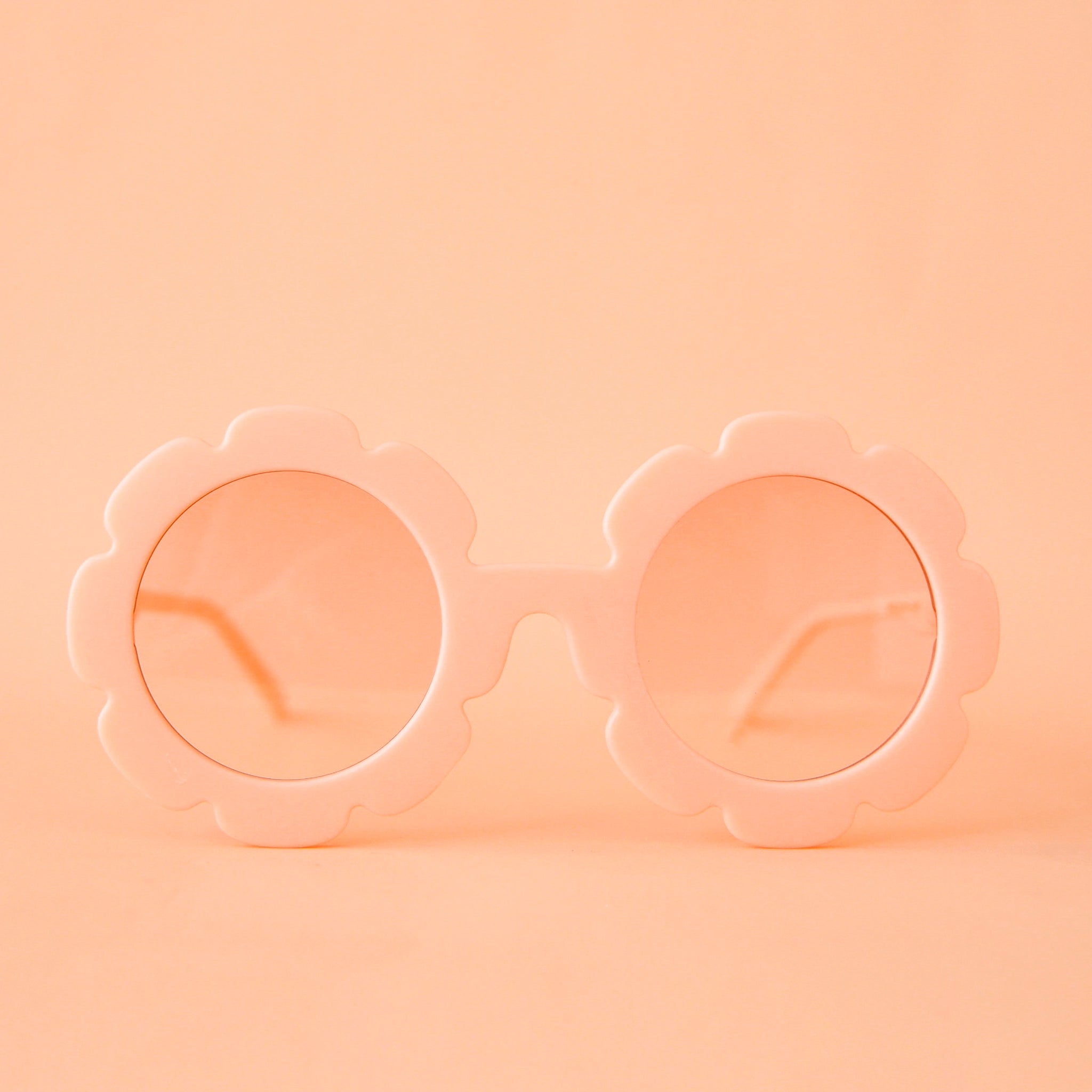 On a peachy background is a pair of flower shaped sunglasses with pink lenses and a apricot colored frame. 