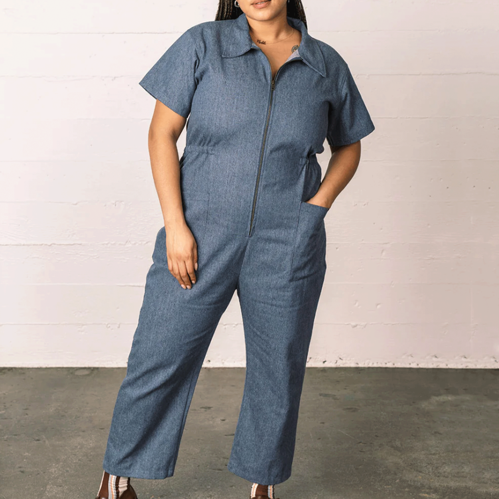 On a neutral background is a model wearing a denim short sleeved jumpsuit with a front zipper and a collar. 