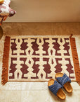 A cream, brown and red geometric patterned bath mat with tassel edges on two sides.