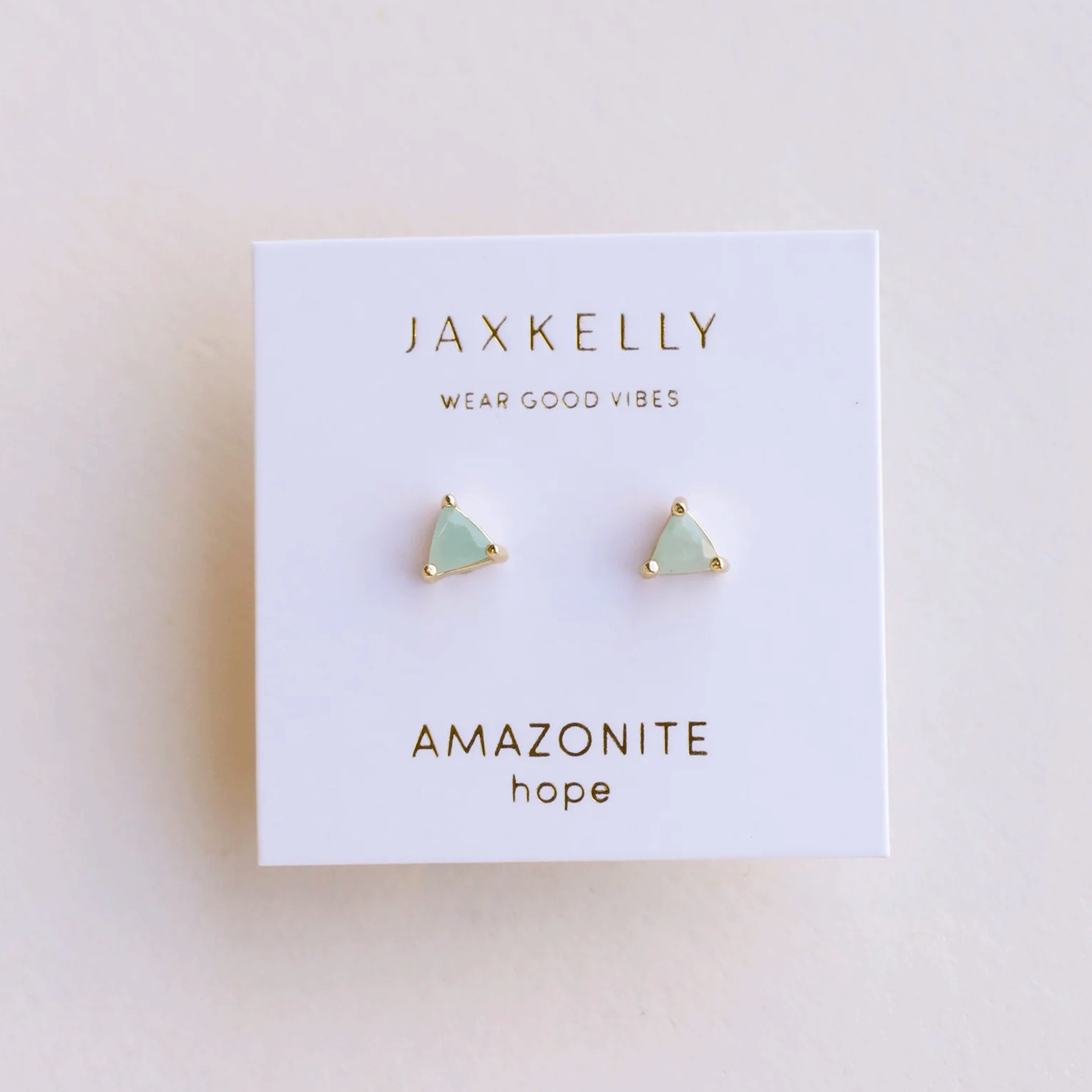 On a cream background is a white square piece of packaging holding a pair of green and gold amazonite stud earrings in a triangle shape along with gold text on the packaging that reads, "JaxKelly Wear Good Vibes Amazonite hope".