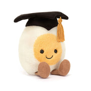 On a white background is a stuffed toy in the shape of a hard boiled egg wearing a graduation cap and smiling. 